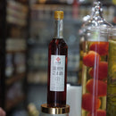 A dark glass bottle with a gold cap and a white Japanese label stands on a clear glass jar with a gold collar, set against a blurred shop background with other jars visible.
